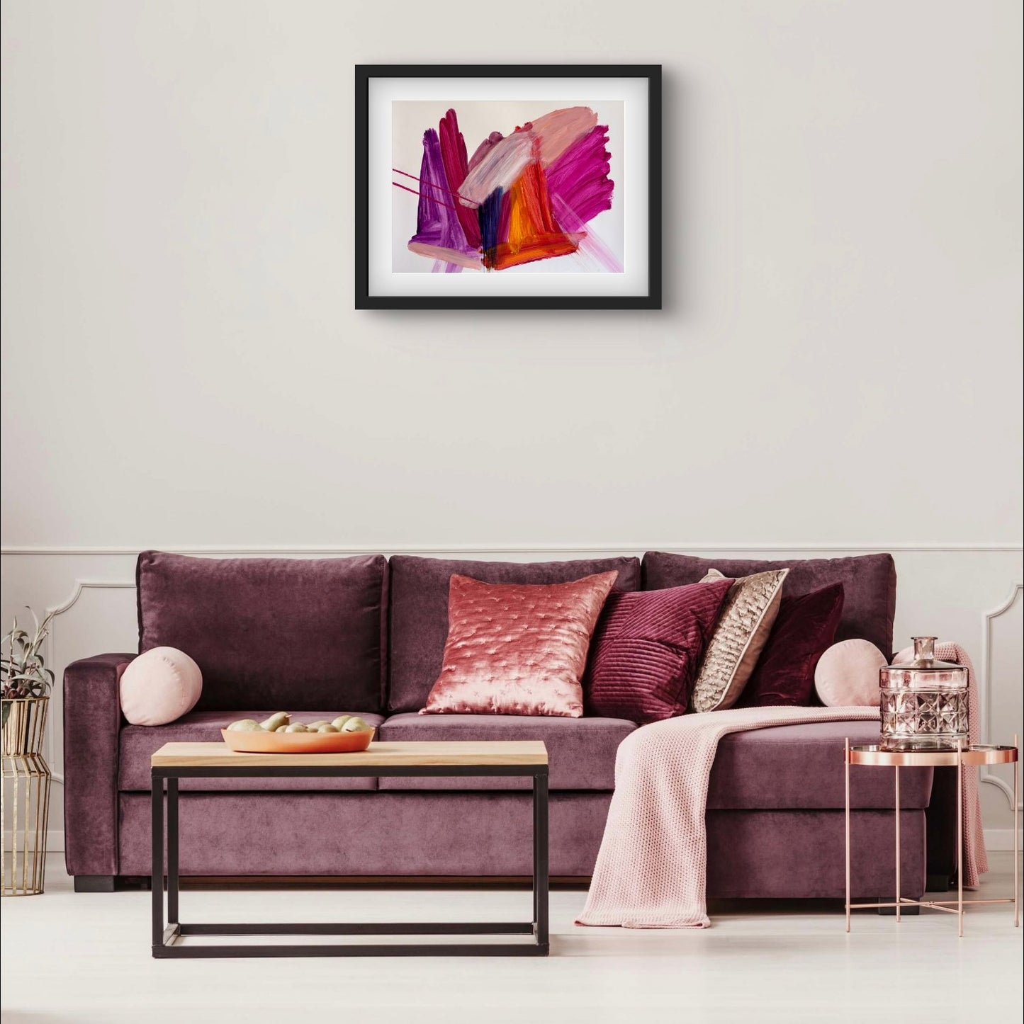 Bright expressive abstract artwork on paper with pink, purple, orange and peach colours in a black frame hanging above a purple couch