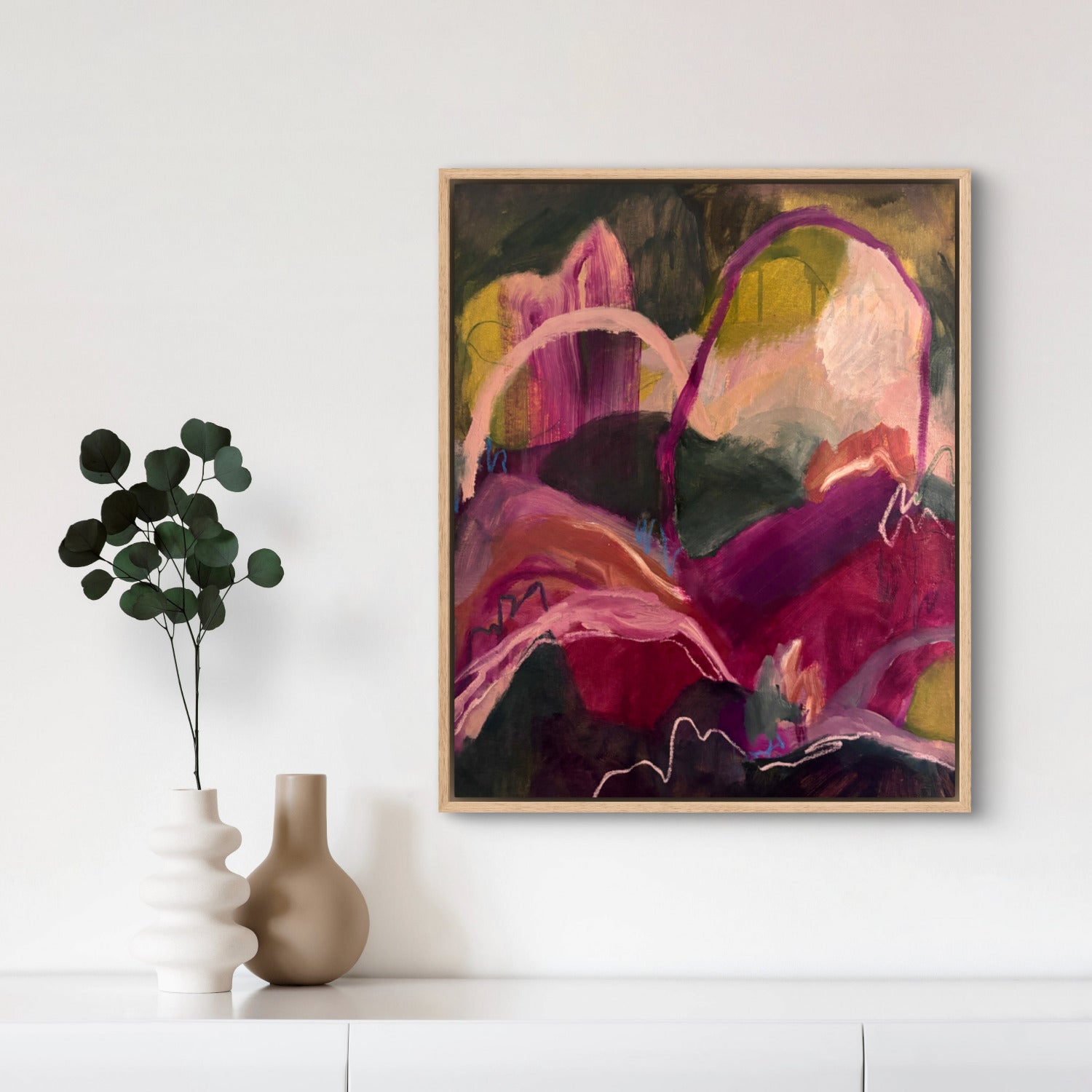 Abstract landscape painting of hills and arches in dark green, magenta and orange, hanging above a white cupboard with two vases