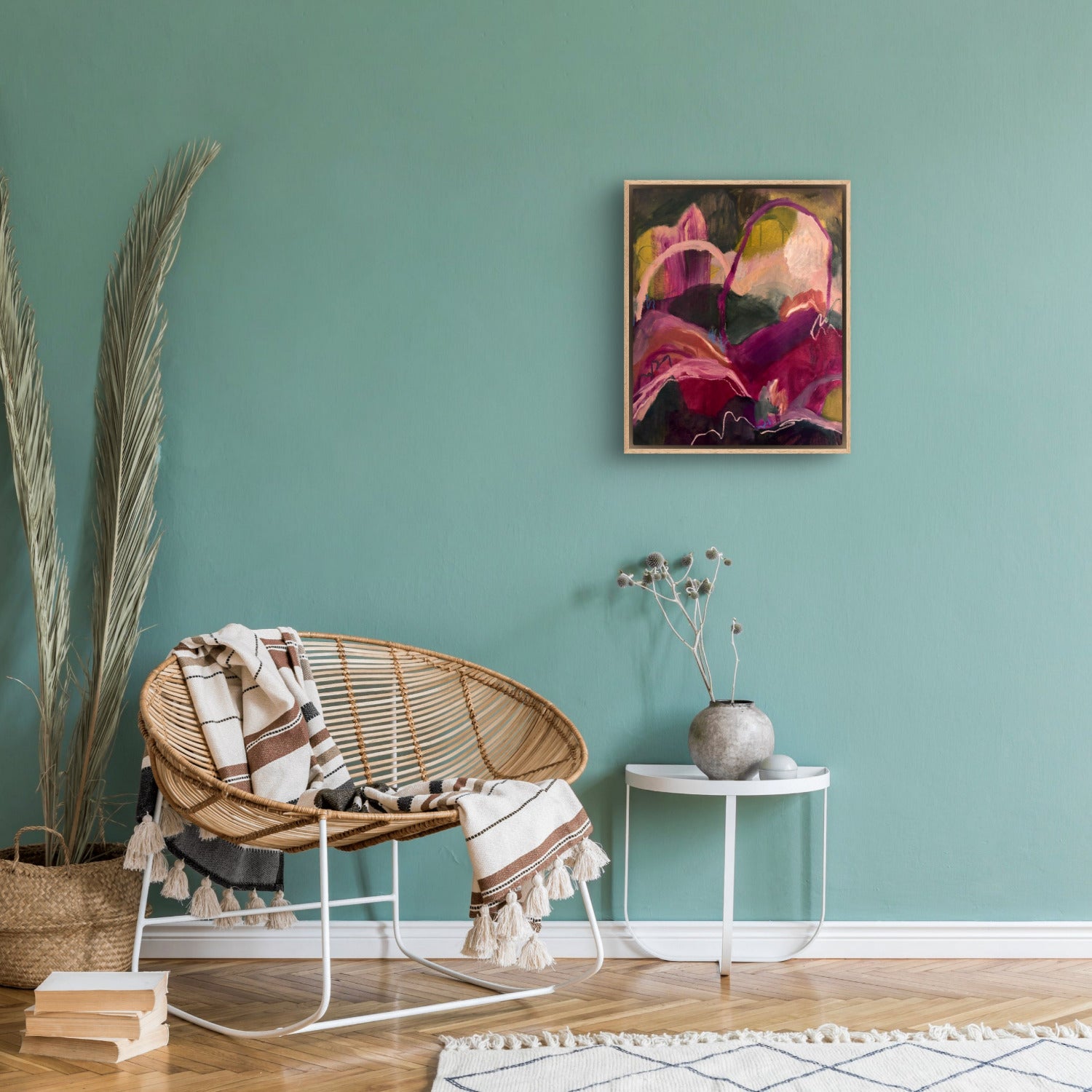 Abstract landscape painting of hills and arches in dark green, magenta and orange, hanging on a sage coloured wall with cane furniture underneath