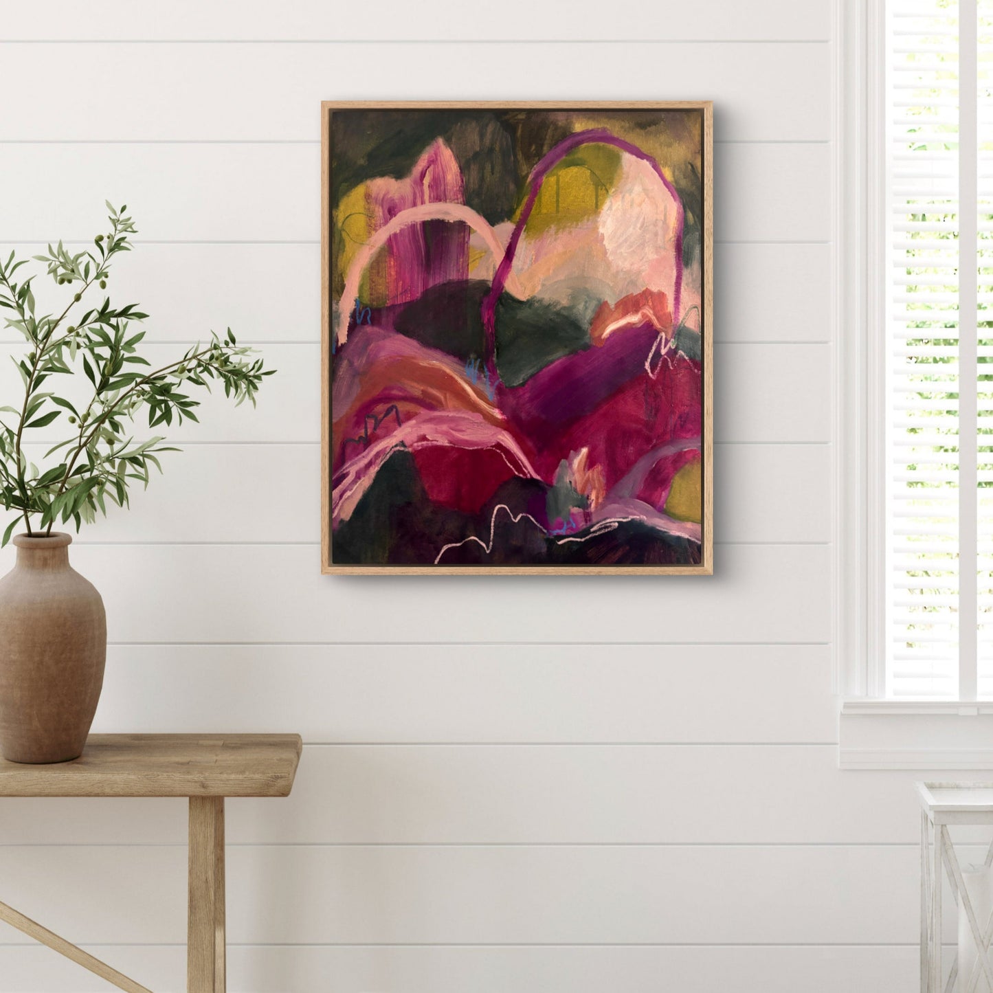 Abstract landscape painting of hills and arches in dark green, magenta and orange, hanging on a white wall with a large vase of green leaves in front