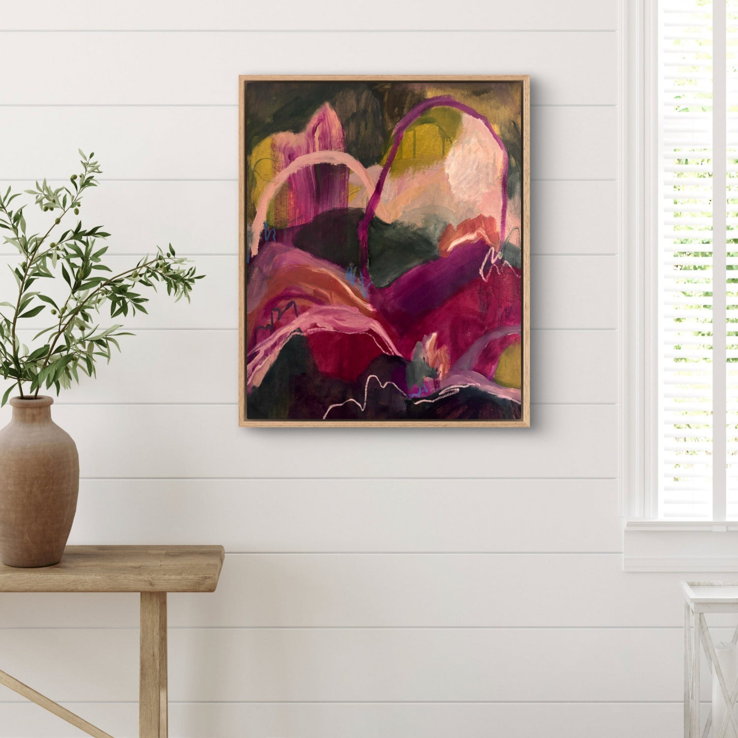 Abstract landscape painting of hills and arches in dark green, magenta and orange, hanging on a white wall with a large vase of green leaves in front