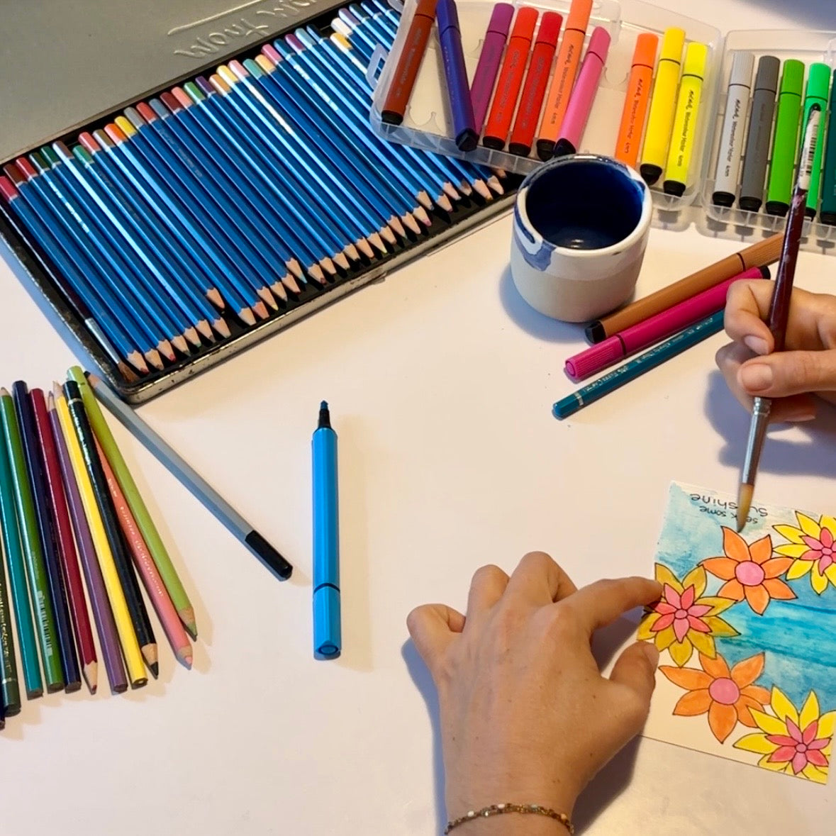 Coloring book with flowers, colored pencils, markers, and a cup on a table.