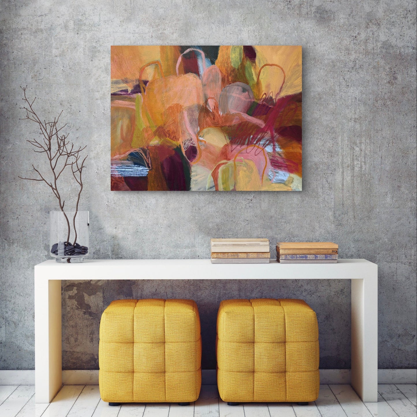 An abstract canvas painting with warm tones of yellow, orange, brown, and red, displayed on a textured gray wall above a white console table with two yellow ottomans.