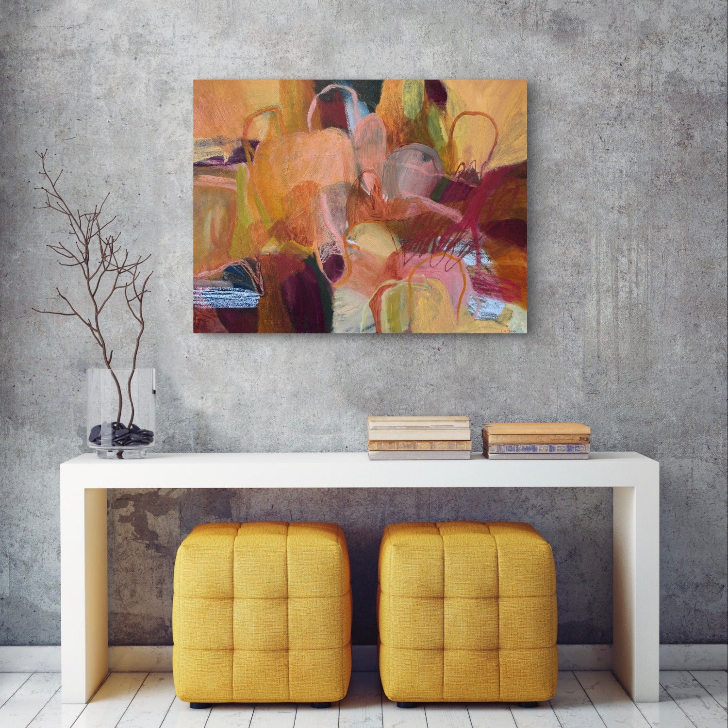 An abstract canvas painting with warm tones of yellow, orange, brown, and red, displayed on a textured gray wall above a white console table with two yellow ottomans.