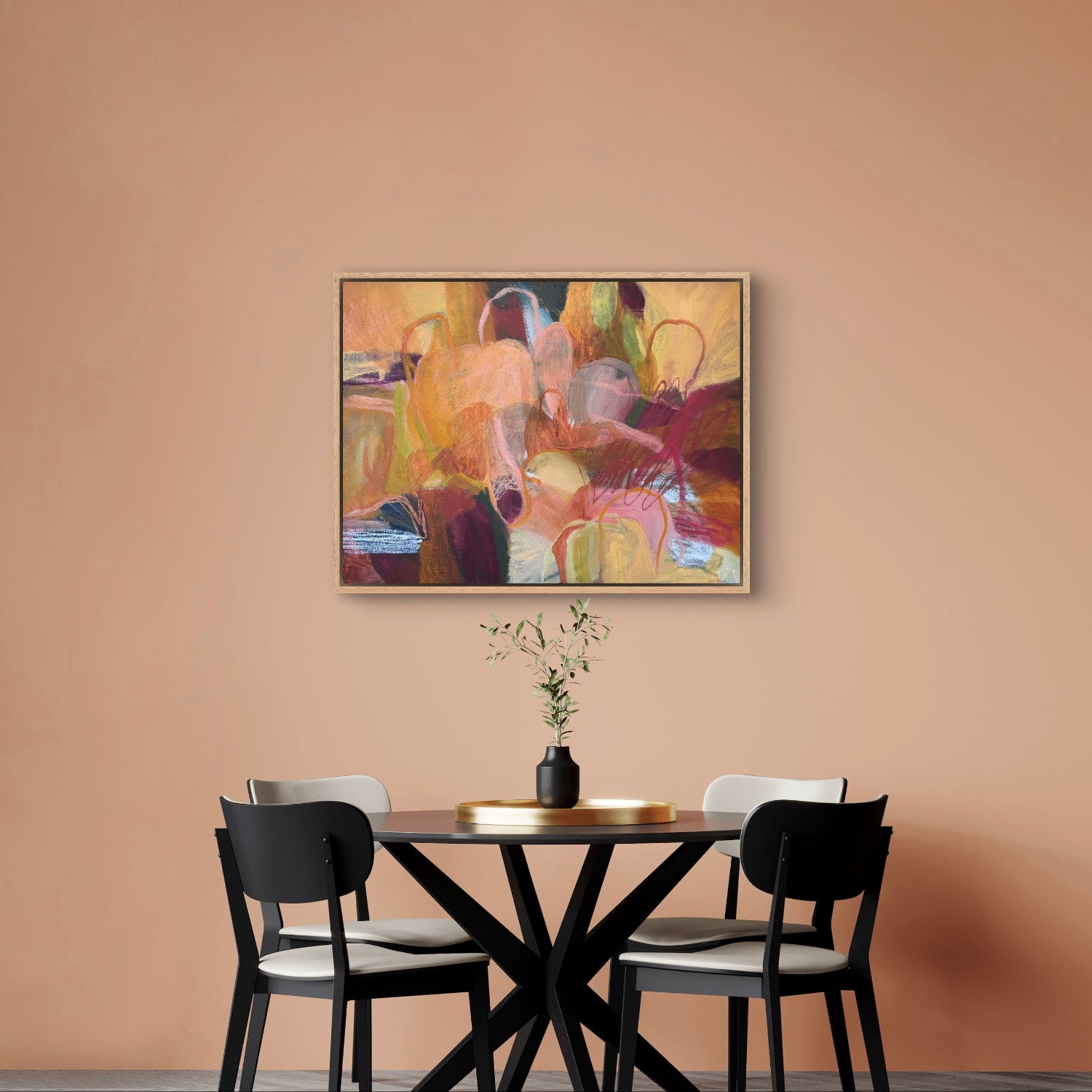 An abstract framed painting with warm tones of orange, yellow, brown, and red hangs on a peach-colored wall above a dining table with chairs.