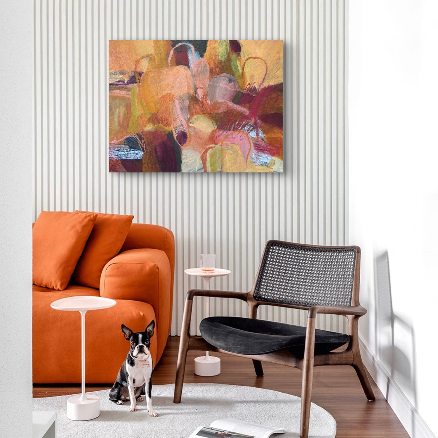 An abstract painting with warm tones of orange, yellow, brown, and red is displayed on a white slatted wall in a modern living room setting with an orange sofa, black armchair, and a small dog.