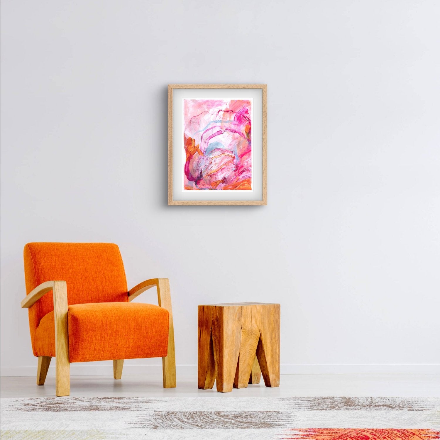 Abstract painting on paper with bright pink, orange and peach colours in an oak frame hanging above a bright orange chair
