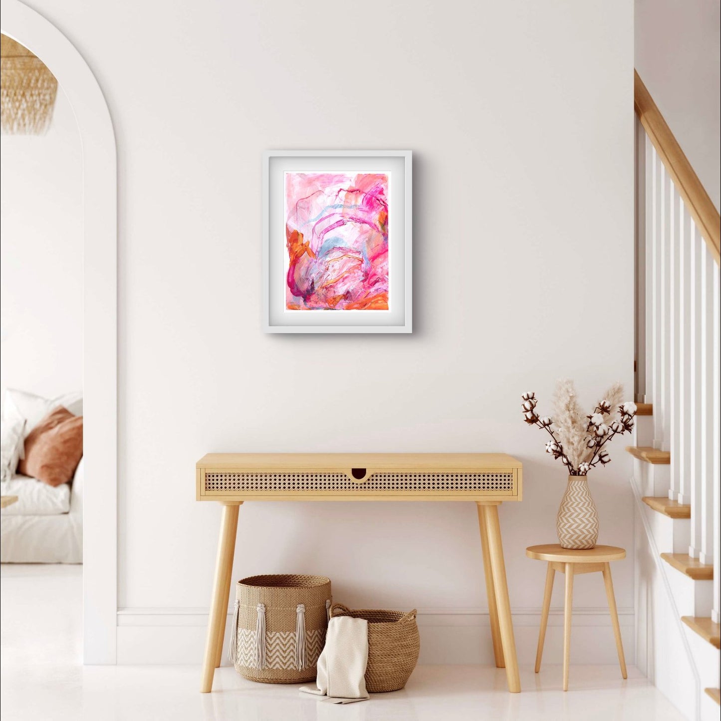 Abstract painting on paper with bright pink, orange and peach colours in a white frame hanging above a console table