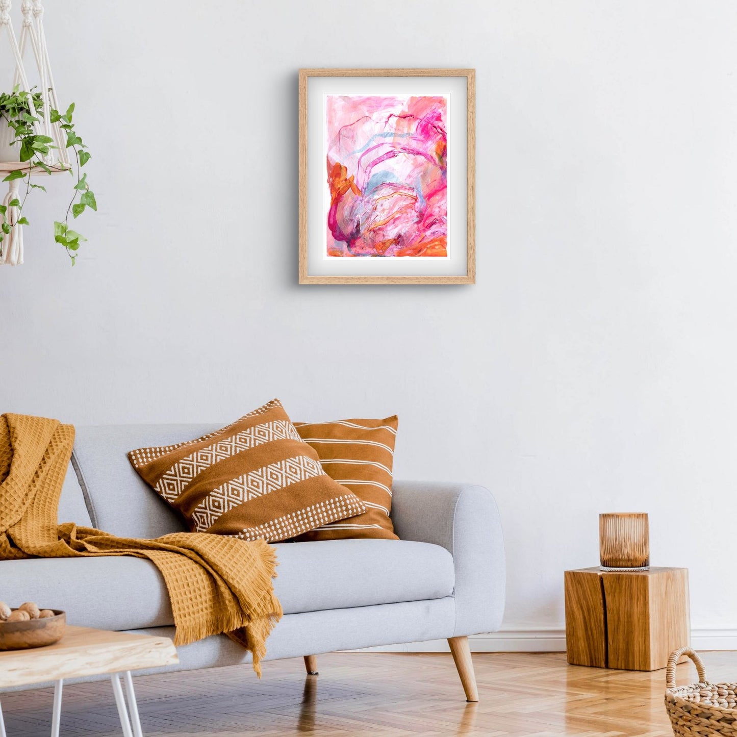 Abstract painting on paper with bright pink, orange and peach colours in an oak frame hanging above a couch with mustard cushions