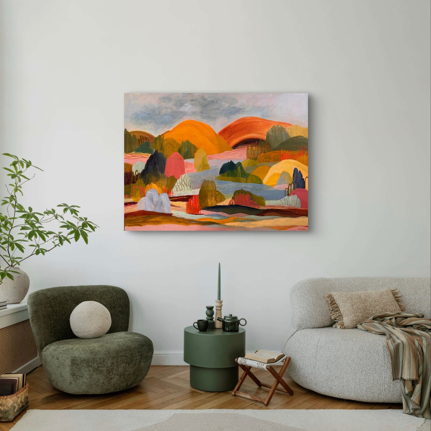 A colorful abstract landscape painting of rolling hills and mountains hangs on a white wall in a modern living room setting with a green armchair, a gray sofa, and a small side table.