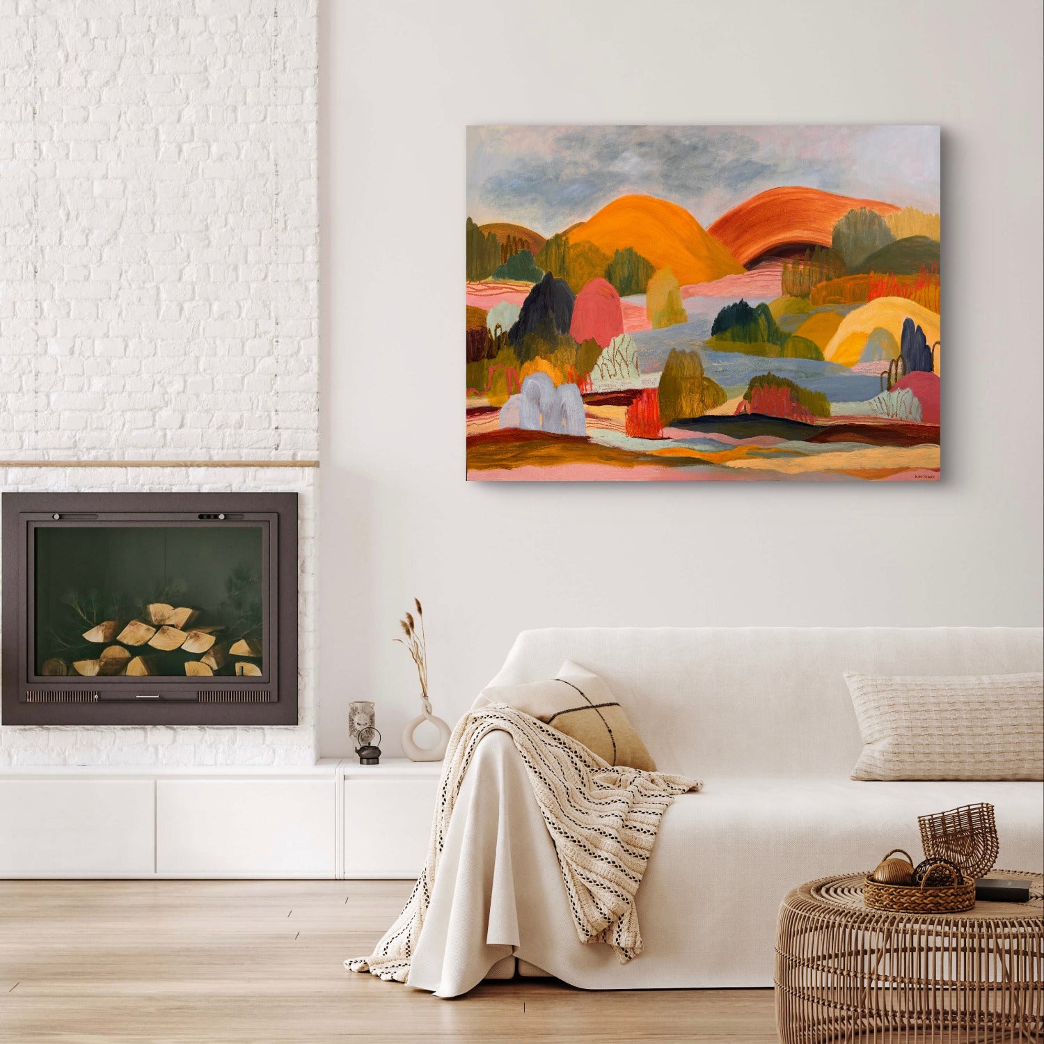A colorful abstract landscape canvas print hangs on a white wall above a white sofa in a modern living room setting with a fireplace and a wicker basket on the floor.
