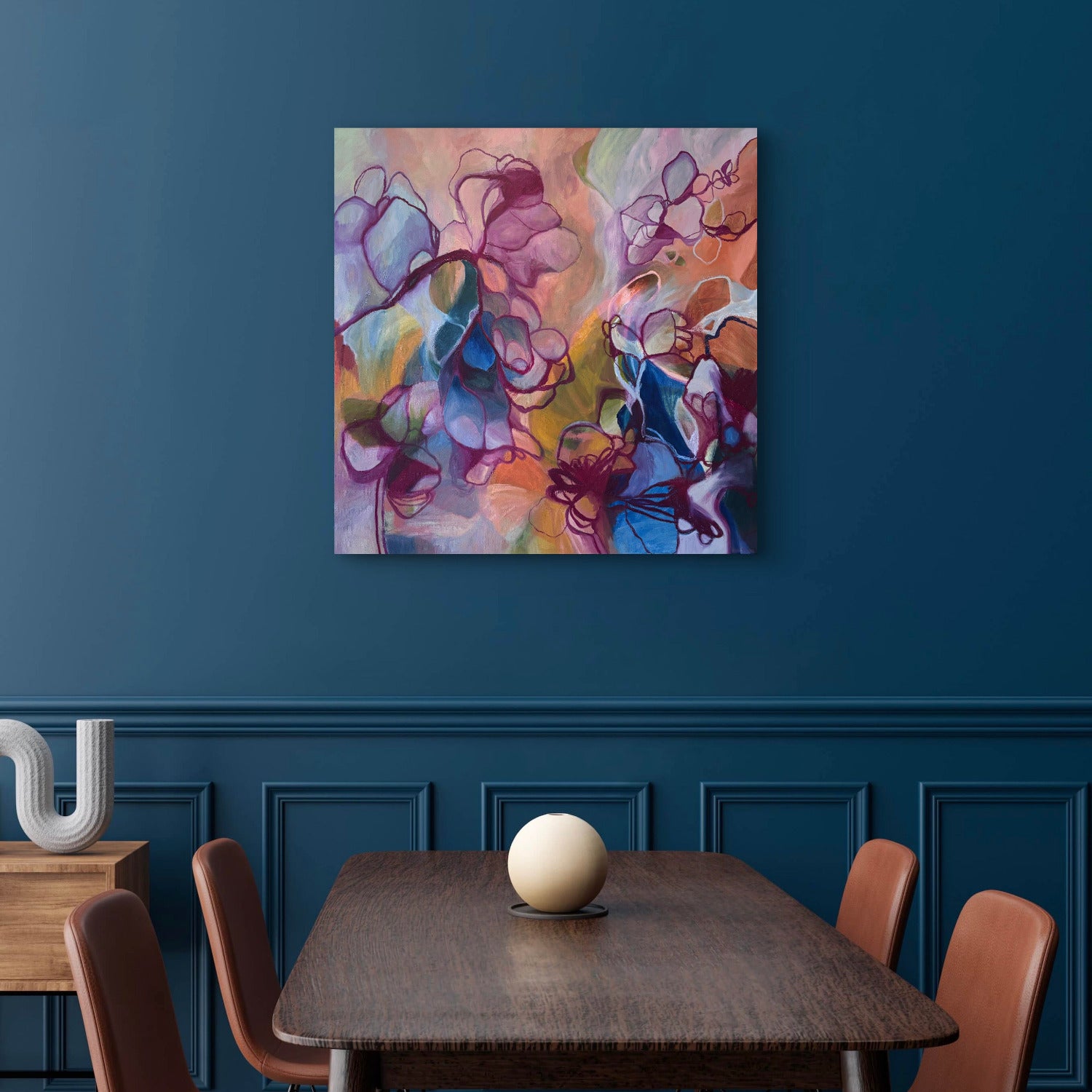 An abstract floral canvas print with purple, blue, pink, and orange tones hanging on a dark blue wall above a wooden dining table with chairs.