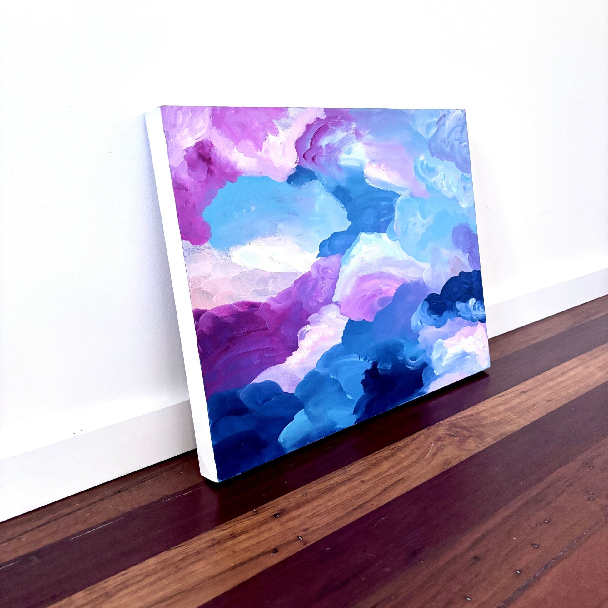 Abstract landscape artwork of clouds with pink,blue and white paint leaning against a white wall on a timber floor