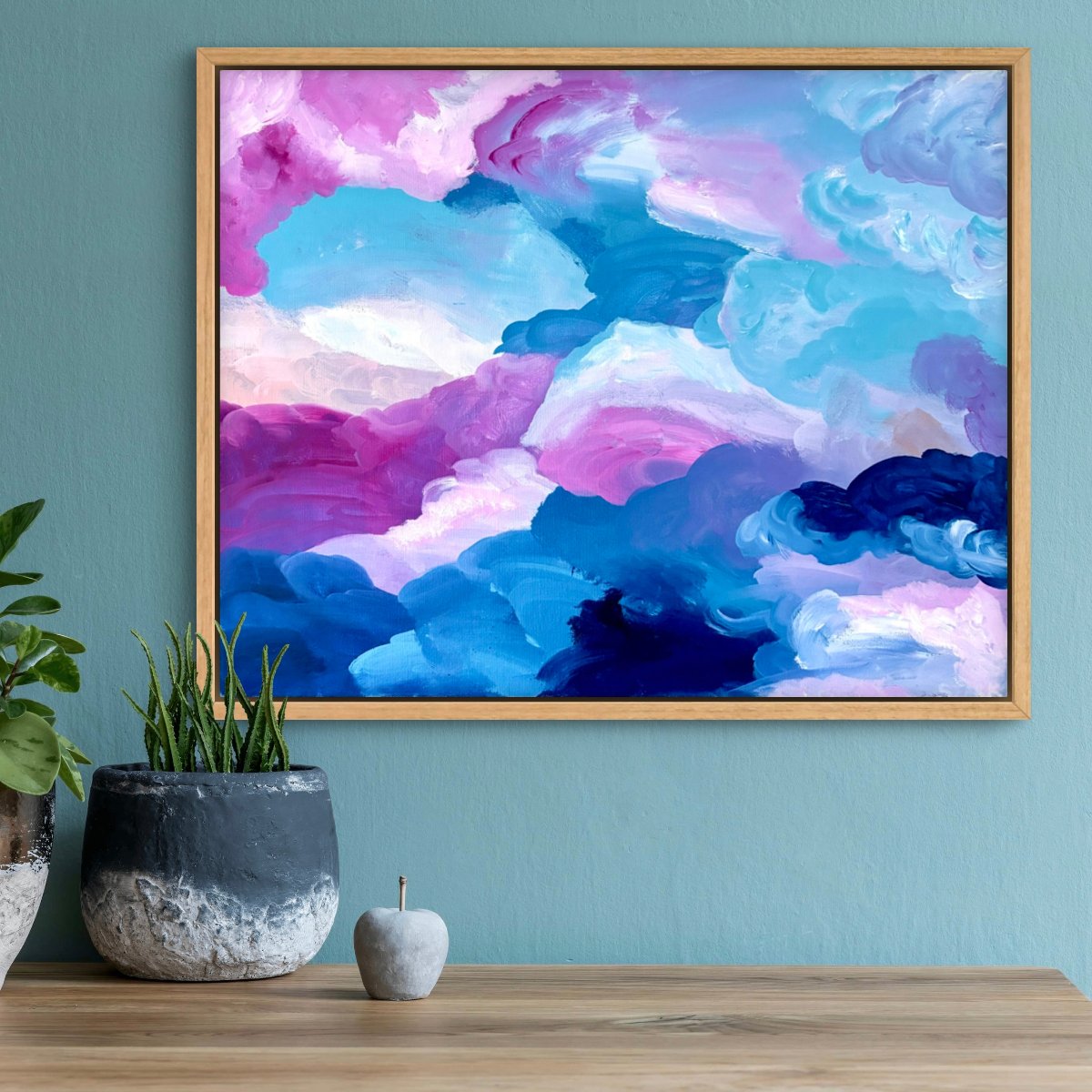 Original paintings of pink and blue clouds framed in an oak frame on a teal wall