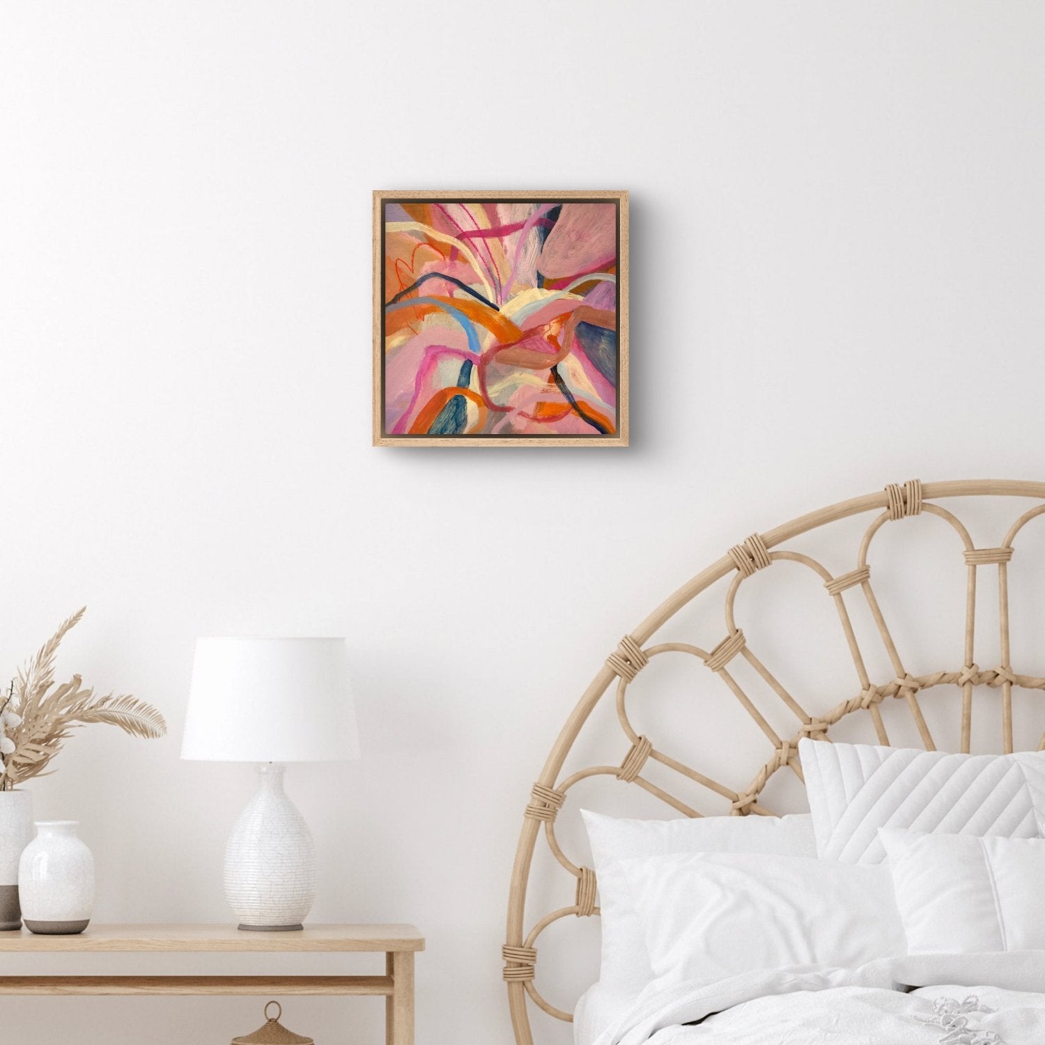abstract acrylic painting with birght pink, orange and blue colours in an oak frame hanging on a white wall above a white and cane bed