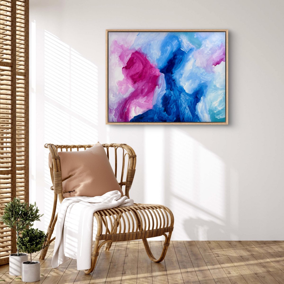 Framed original abstract painting with blue, pink and white colours hanging on a wall with a cane chair in front