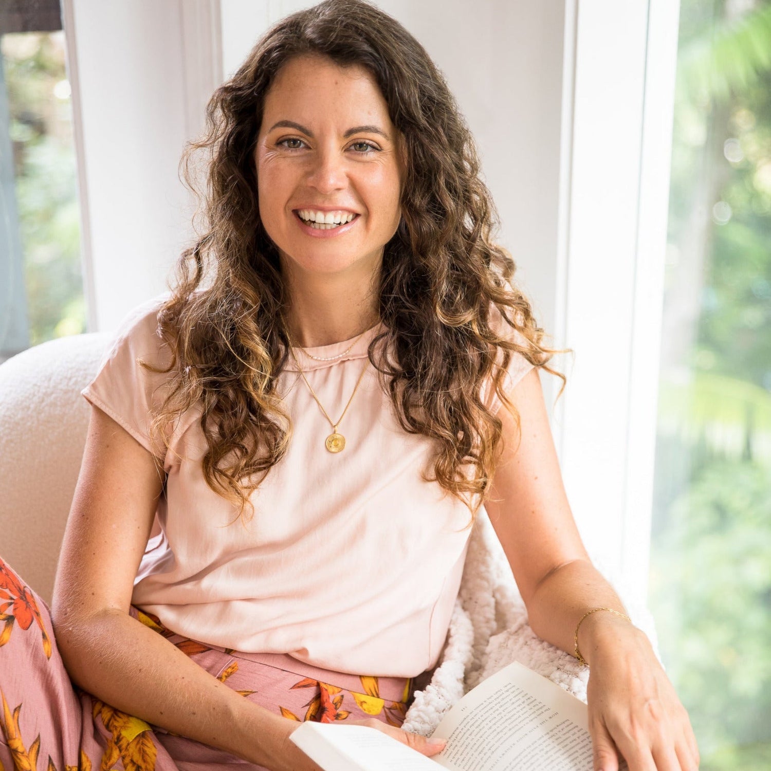 Woman sitting on a couch holding a book, smiling at the camera.