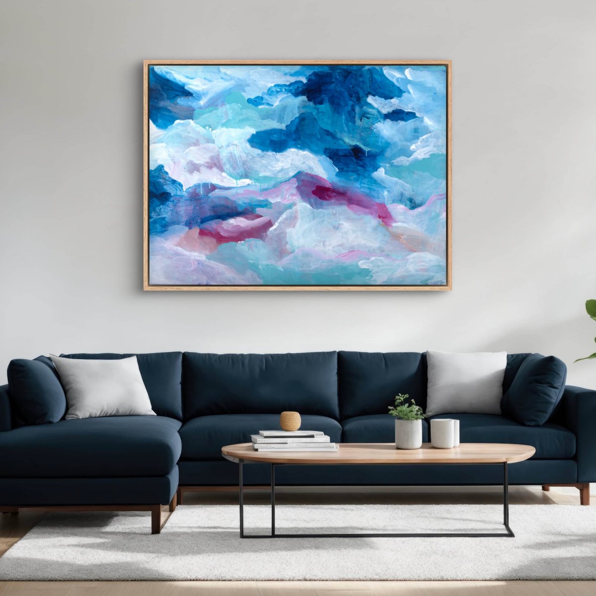 Original painting of abstract clouds in blue, white and pink in an oak frame hanging on a white wall with a dark teal couch in front