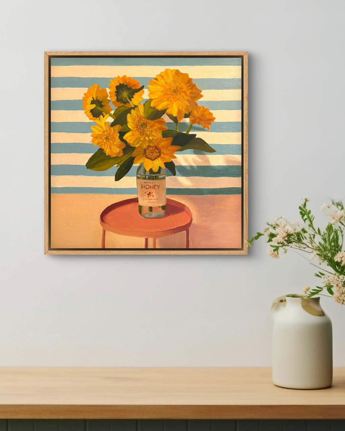 Framed artwork of sunflowers on a striped background, displayed on a wall above a shelf with a vase.