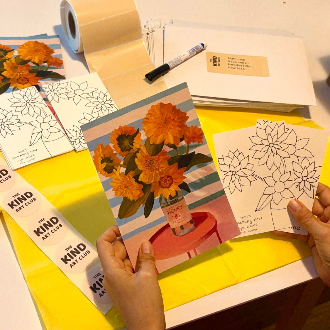 Table with art supplies, coloring books, and a person holding a card with a floral design.